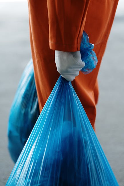 A close-up of a person wearing orange disposable overalls and white work gloves, holding a large, tightly knotted blue plastic rubbish bag at their side. The scene suggests an outdoor setting, possibly on a concrete or asphalt surface, with a blurred background that appears to be a paved or industrial area. The individual is preparing to transport waste, indicative of rubbish removal activity, with the orange protective gear providing visibility and safety during handling. The blue bag, made of translucent plastic material, reveals some irregular shapes inside, hinting at mixed waste materials. This visual context aligns with private waste collection or alternative rubbish disposal services, like those offered by Rubbish Removal Watford, especially when handling waste on-site or in non-public collection scenarios. The lighting is natural, even, and unobstructed, emphasizing clear visibility of the waste packaging and safety equipment used in rubbish removal processes.