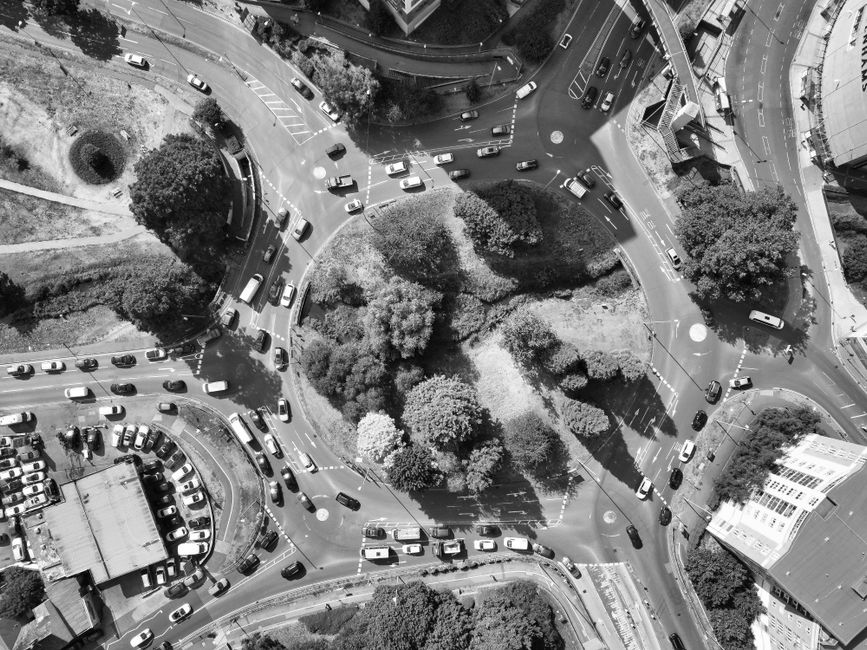 A high-altitude aerial view of a roundabout surrounded by multiple lanes of traffic with numerous cars navigating the intersection. In the center of the roundabout, there is a landscaped island densely populated with trees and shrubs, featuring various textures from leafy canopies to more open grassy areas. The trees vary in size and shape, with some rounded and others more irregular, casting shadows onto the surrounding pavement. To the lower left, there is a large parking lot filled with parked vehicles, bordered by a building with a flat roof. Adjacent to the road network, a few trees and patches of greenery provide additional natural elements within the urban setting. The roads are asphalt with clearly marked lane lines and roundabout indicators, and the scene is illuminated by natural daylight, creating sharp contrasts between light and shadow. This snapshot illustrates a typical urban traffic management area, highlighting the importance of organized vehicle flow near busy stations, such as Watford Junction, and how private waste removal services like Rubbish Removal Watford might operate in proximity to such busy transport hubs, focusing on efficient on-site clearance and non-infrastructure related waste handling.