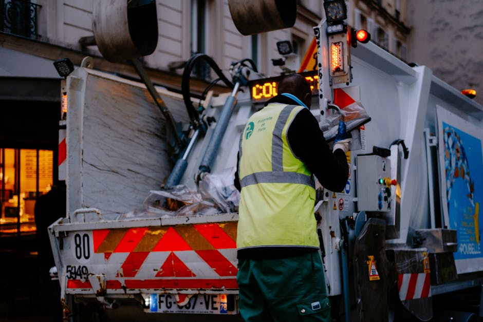 A close-up of a person wearing orange disposable overalls and white work gloves, holding a large, tightly knotted blue plastic rubbish bag at their side. The scene suggests an outdoor setting, possibly on a concrete or asphalt surface, with a blurred background that appears to be a paved or industrial area. The individual is preparing to transport waste, indicative of rubbish removal activity, with the orange protective gear providing visibility and safety during handling. The blue bag, made of translucent plastic material, reveals some irregular shapes inside, hinting at mixed waste materials. This visual context aligns with private waste collection or alternative rubbish disposal services, like those offered by Rubbish Removal Watford, especially when handling waste on-site or in non-public collection scenarios. The lighting is natural, even, and unobstructed, emphasizing clear visibility of the waste packaging and safety equipment used in rubbish removal processes.