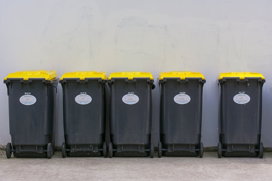 Five large wheeled rubbish bins lined up against a plain, light gray wall, with yellow lids and black bodies made of durable plastic. Each bin has a white oval label on the front displaying identification numbers and company details. The bins are positioned on a flat concrete surface, with some slight scuff marks visible at their bases. The scene appears to be outdoors or in an open area designated for waste collection, with natural or diffuse lighting illuminating the scene evenly. The orderly arrangement suggests they are intended for domestic or commercial waste disposal, aligning with private rubbish removal or on-site clearance services offered by Rubbish Removal Watford. The uniform appearance emphasizes cleanliness and organization, with the bins ready for collection or servicing as part of scheduled waste management activities, highlighting the practical aspects of effective rubbish management and the importance of proper waste handling outside of municipal systems.