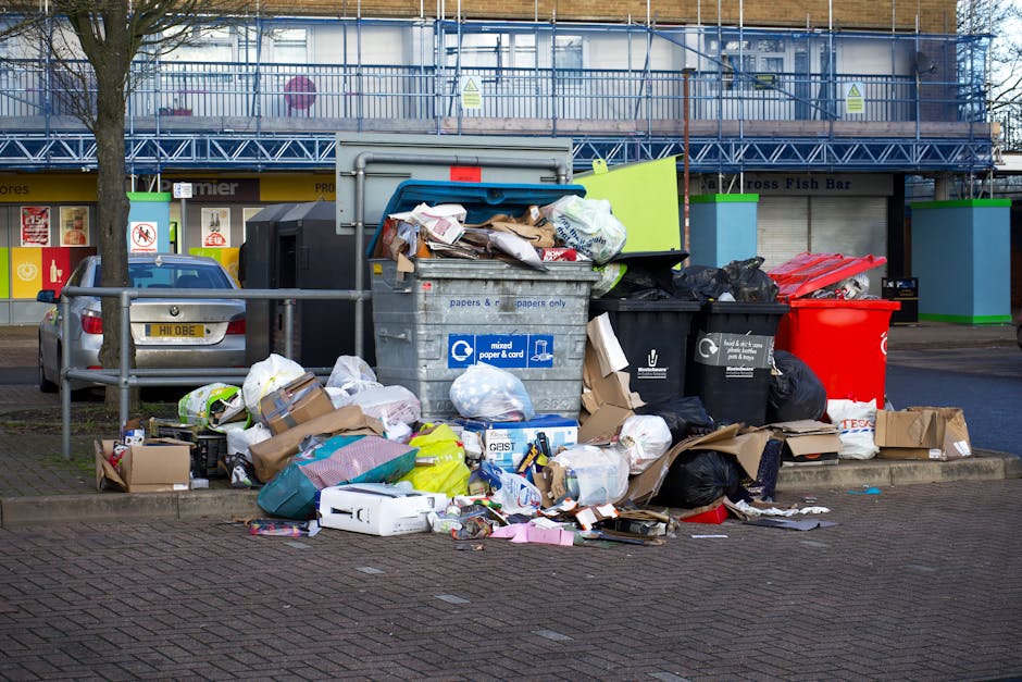 The image depicts a large pile of mixed waste and rubbish bags overflowing from multiple recycling and trash bins situated on a paved sidewalk area outside a commercial building. The waste includes various items such as flattened cardboard boxes, plastic bags, paper debris, and packaging materials, some of which are spilling onto the ground around the bins. The bins are of different colours and materials, including a blue bin labeled for mixed paper and card, a black bin, and a red bin. Behind the waste collection point, there is a metal railing separating the pavement from parked vehicles, with one visible silver car on the left side. The backdrop features a commercial storefront with signage, a blue scaffold or construction framework on the upper level of the building, and trees partially visible at the top. The scene appears to be in an urban or shopping area, illustrating a typical instance of excess rubbish accumulation, which could potentially be addressed through private waste removal services such as those offered by Rubbish Removal Watford, especially for on-site clearance or alternative disposal arrangements outside regular local authority collection.