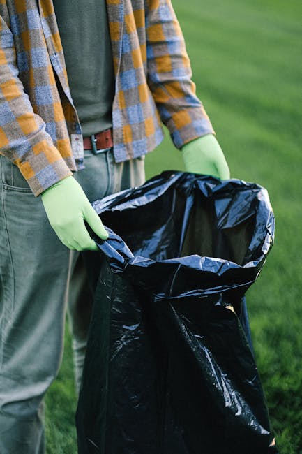 A person wearing a green long-sleeved shirt, a yellow and gray checkered jacket, and light gray pants, standing outdoors on a grassy area, is holding a large black plastic rubbish bag open with both hands, wearing lime green gloves. The black rubbish bag is partially open and appears empty, ready for waste collection. The background shows a well-maintained lawn with blurred green grass and possibly some natural surroundings. The scene suggests an outdoor rubbish collection or waste disposal activity, potentially conducted by Rubbish Removal Watford or a similar waste management service, emphasizing private, on-site rubbish handling. The lighting indicates natural daylight, providing clear visibility of the scene, with no visible vehicles or additional equipment present. This image exemplifies individual or community-led rubbish removal efforts, suitable for environmental cleanup or waste management initiatives.