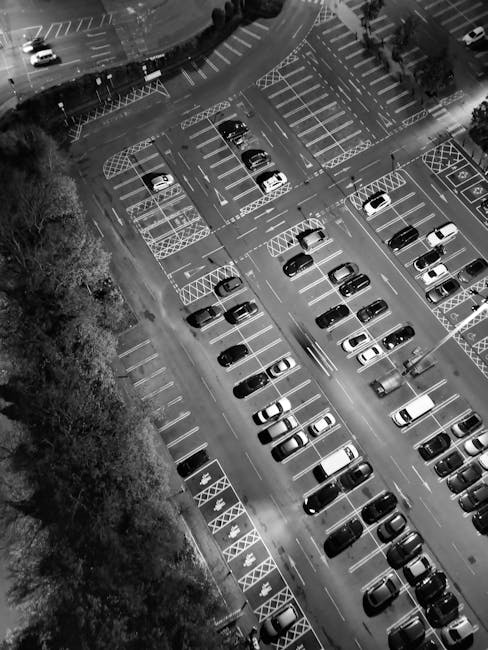 An aerial view of a large outdoor parking lot during nighttime, showing multiple rows of parked vehicles including sedans, hatchbacks, and SUVs in various shades of black, white, and grey. The parking area is divided by marked parking bays, with some bays reserved or designated with crosshatched white lines. Surrounding the parking lot, there are trees on the left side with leafy foliage visible in the dim lighting. The lot features metal barriers and markings indicating pedestrian walkways, and some vehicles are positioned near the edges of the parking bays. The overall scene has limited illumination, with shadows cast by the vehicles and nearby structures, giving a clear impression of a busy, organized parking area potentially used for daily access or alternative vehicle storage, as could be associated with private or commercial waste management companies like Rubbish Removal Watford. This scene subtly underscores the importance of organized vehicle management in support of effective rubbish and waste collection operations by independent service providers.